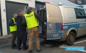 Jeden z policjantów otwiera drzwi radiowozu , drugi stoi z zatrzymanym