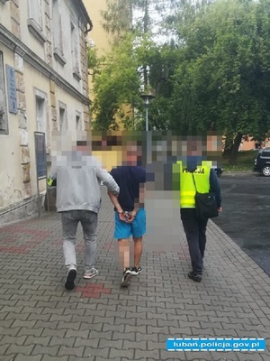 dwóch nieumundurowanych policjantów prowadzi osobę zatrzymaną do przestępstwa. jeden z policjantów posiada kamizelkę odblaskową żółtą z napisem POLICJA