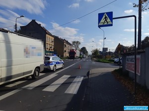 jezdnia z przejściem dla pieszych, na przejściu stoi biały bus, obok oznakowany radiowóz policyjny, w oddali widoczny policjant kierujący ruchem na drodze