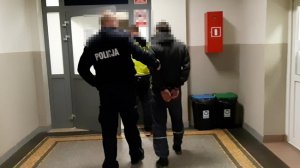 Na zdjęciu widać dwóch policjantów jeden w umundurowaniu a drugi po cywilnemu w kamizelce z napisem Policja otwierają drzwi do pomieszczenia wchodzą wraz z zatrzymanym mężczyzną.