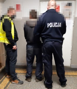 Zdjęcie przedstawia policjanta w umundurowaniu służbowym oraz policjanta w kamizelce żółtej oraz mężczyznę zatrzymanego do przestępstwa